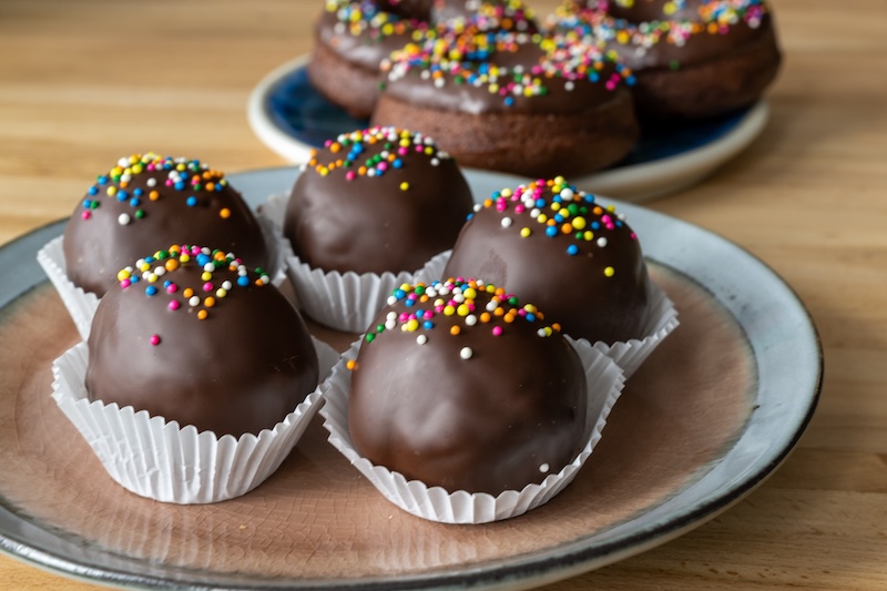 A small round plate of round double chocolate covered truffle balls with colorful sprinkles next to a blue plate with three chocolate glazed baked donuts. The sweets are on a wooden oak table.