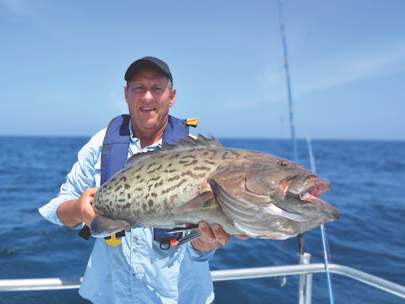 Man holding a Gag Grouper on a fishing boat in the ocean with a fishing pole behind him