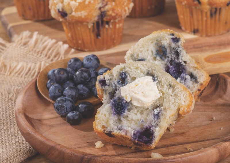 Sliced blueberry muffin and spoonful of berries on a wooden plate with muffins in background