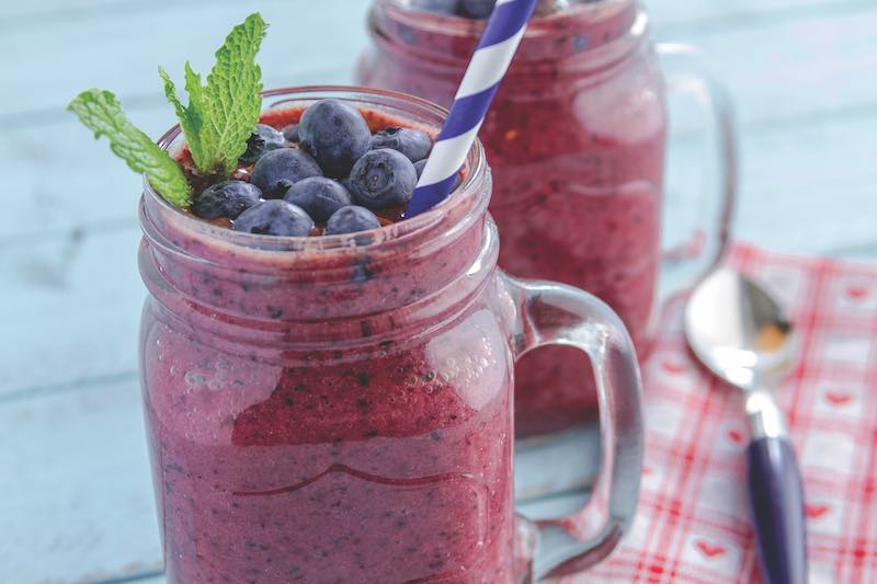 Close up of 2 Mason jars filled with blueberry and blackberry fresh fruit smoothie sitting on blue wood background with straws and heart napkin