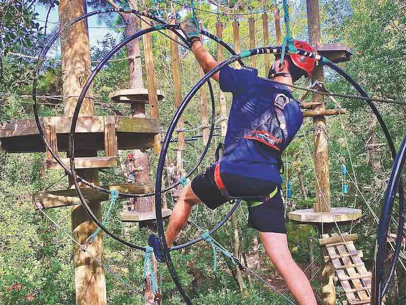 Person wearing a helmet and harness completing an obstacle course in the air with trees in the background