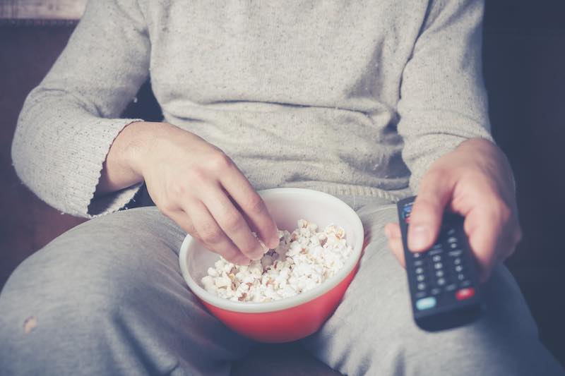 Young man is sitting on a sofa and eating popcorn while watching television
