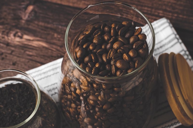 Storage of coffee and tea in glass jars. Glass jars with wooden lids filled with coffee beans and tea leaves stand on a table against a dark background.