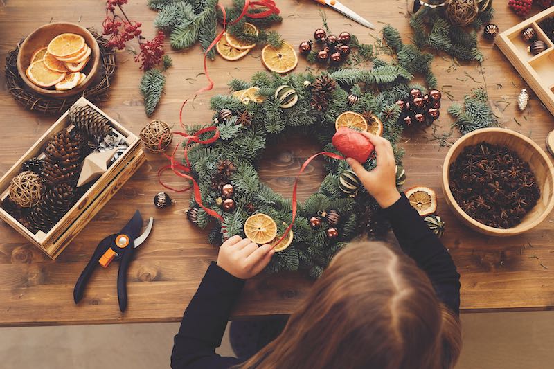 High angle view of busy woman standing at desk and using decorative ribbon while making Christmas wreath