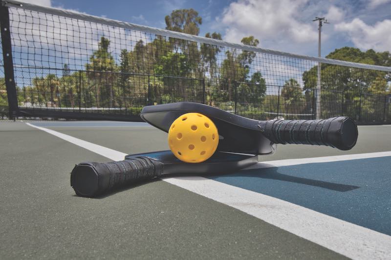 Closeup of a pickleball and paddles on an empty court under sunny skies.