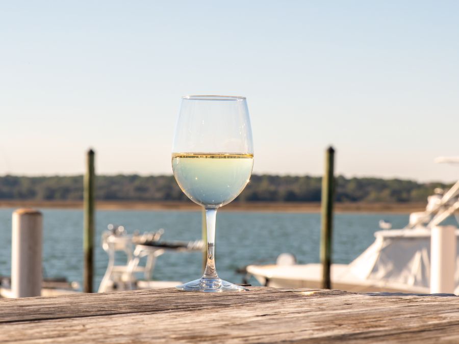 A glass of white wine sitting on a dock with boats and water in the background