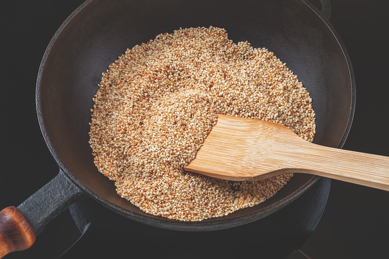 Toasting sesame seeds in a cast iron skillet. Organic benne grains roasting in a frying pan. White til as asian cuisine ingredient. Sesamum indicum as calcium source for healthy eating. Top view.