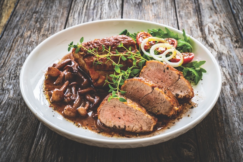 Roasted pork loin with shimeji mushrooms in sauce, fresh thyme, greens and cherry tomatoes on wooden table