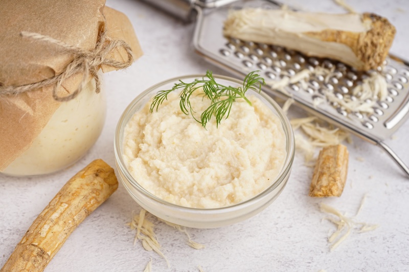 Jar and bowl of horseradish sauce with horseradish roots on white background