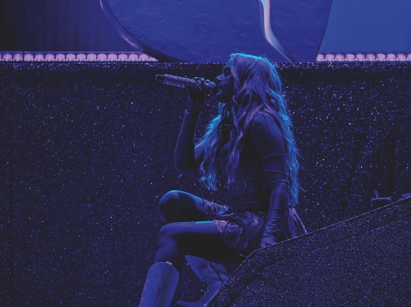 Megan Moroney sitting on stage singing into microphone with dark purple/blue stage lighting