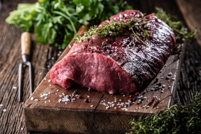Raw rib eye steak on a cutting board with herbs in the background