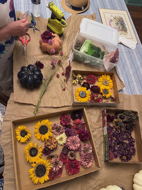 Dried sunflowers and red and pink flowers sit on a table with faux pumpkins and other craft supplies