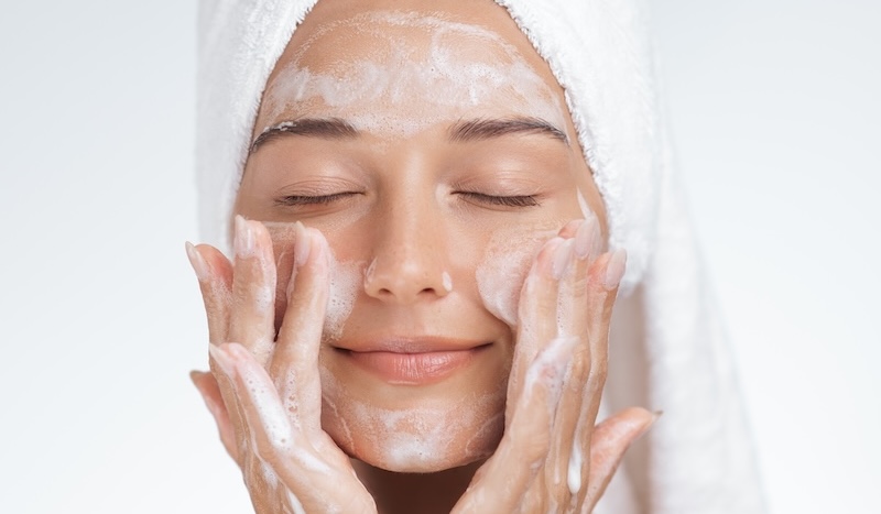 Caucasian woman with eyes closed and towel on head cleansing her face with foam. High quality skincare and spa image perfect for cosmetics, facial care, wellness or beauty promotional design.