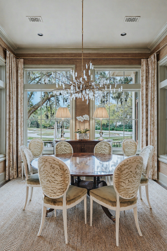 Dining room with a round table, chandelier and tall windows overlooking the landscape