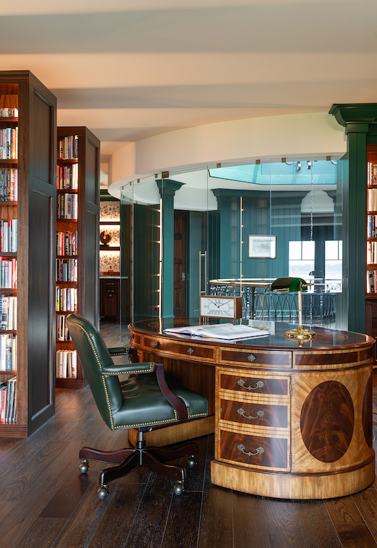Home library with floor-to-ceiling dark wood bookcases, a green leather chair, curved wood desk and glass interior wall