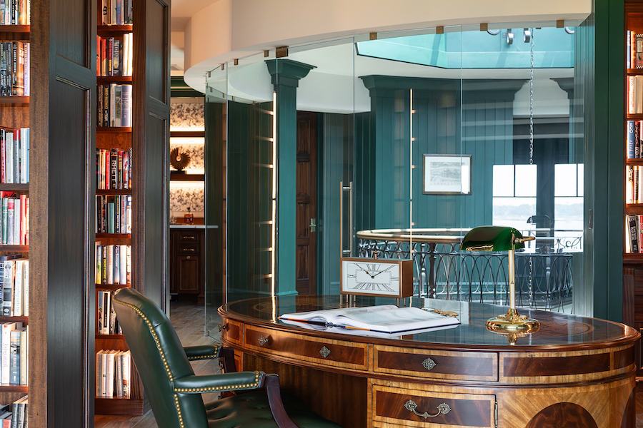 Home library with floor-to-ceiling dark wood bookcases, a green leather chair, curved wood desk and glass interior wall