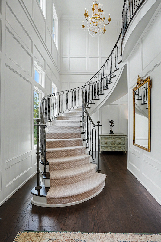 Grand curved staircase with dark metal railing rising through a white paneled foyer beneath a crystal chandelier