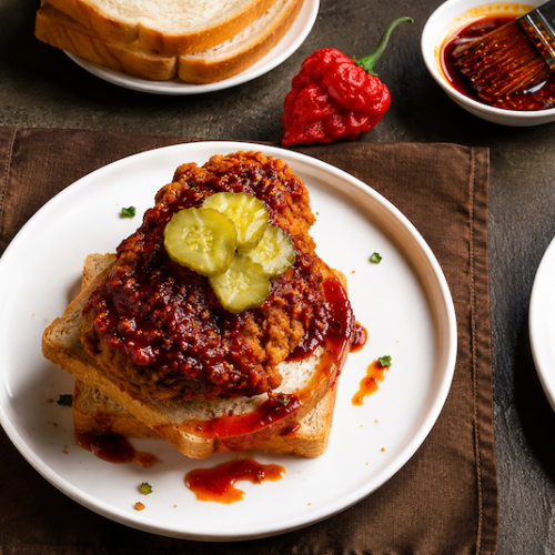 Glazed fried chicken topped with pickle chips and served on white bread, with red peppers nearby.