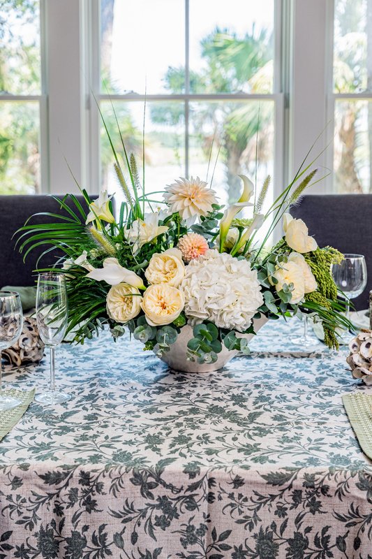 White hydrangea, cream roses, dahlias, calla lilies and greenery arranged in a pale vessel on a patterned tablecloth