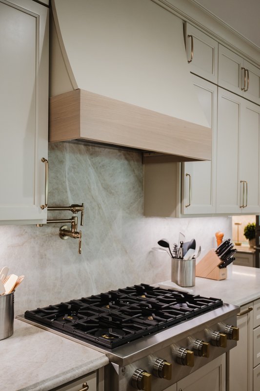 Kitchen range with stone backsplash, pale wood hood and brass pot filler
