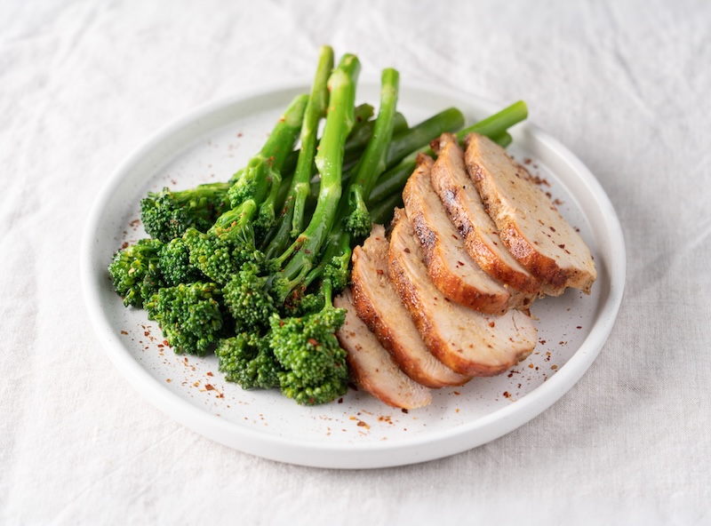 Cooked broccolini served alongside sliced roasted meat on a white plate.