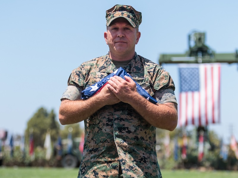 Man wearing camouflage clothing and a baseball cap holds a small object while standing outdoors.