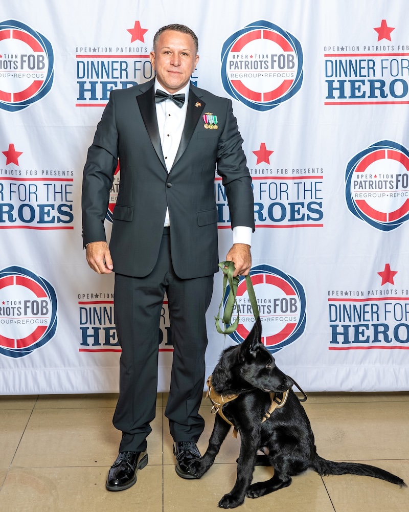 TJ Johnston stands in formal attire beside a black service dog at an Operation Patriots FOB Dinner for the Heroes event.