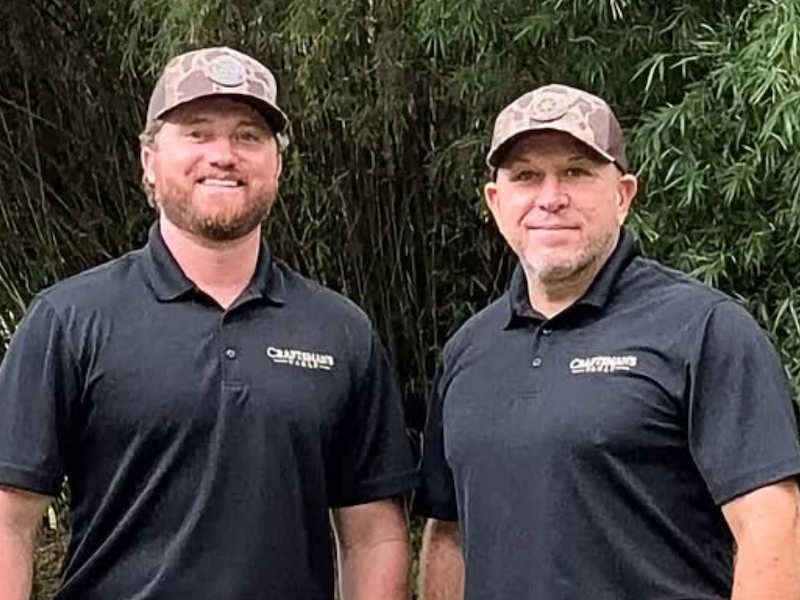 TJ Johnston and Malcolm Claxton stand outdoors wearing matching black Craftsman’s Vault Construction polo shirts.