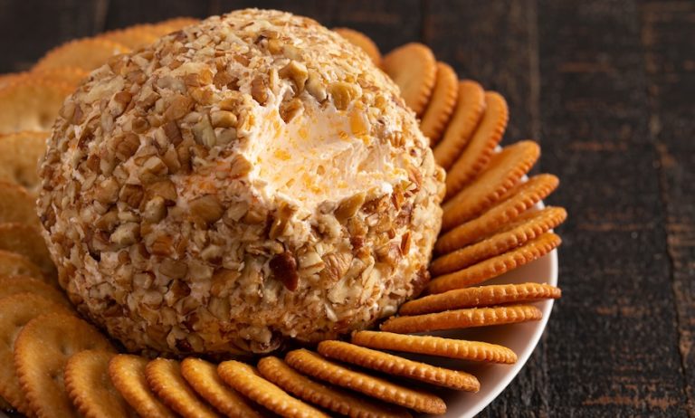 Cheese ball coated in chopped nuts and surrounded by crackers on a serving plate.