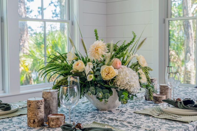 White hydrangea, cream roses, dahlias, calla lilies and greenery arranged in a pale vessel on a patterned tablecloth