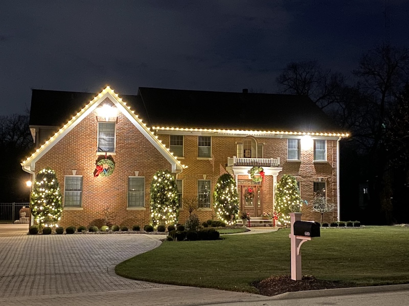 Two-story brick home with warm white Christmas lights outlining the roof and illuminating the front shrubs.