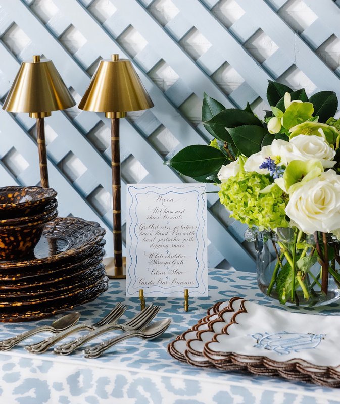 Blue-and-white patterned tablecloth with stacked plates, silverware, flowers and two small gold lamps