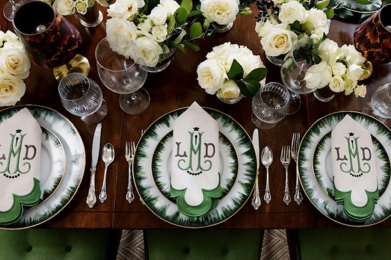 Overhead view of a wooden dining table with green-and-white monogrammed plates, white roses, glassware and silverware