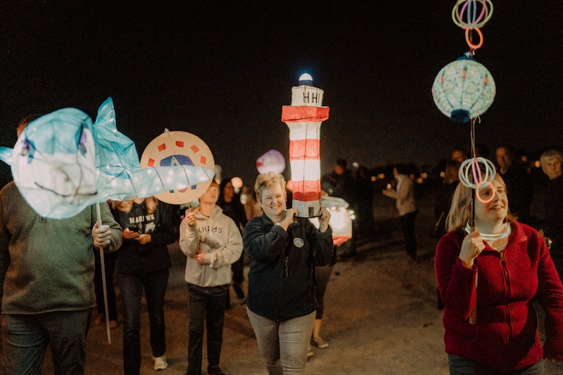 Participants carry illuminated handmade lanterns along a beach at night.
