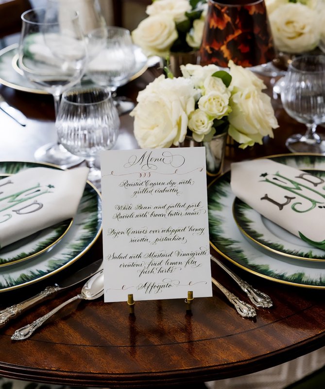 Printed menu card on a wooden dining table surrounded by white flowers, patterned plates, glasses and silverware
