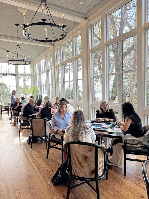 Groups of people playing mahjong at tables in a bright room with tall windows