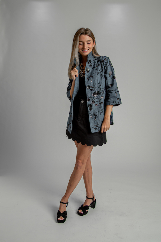 Woman wearing a textured blue jacket, black top, black scalloped skirt and black heels.