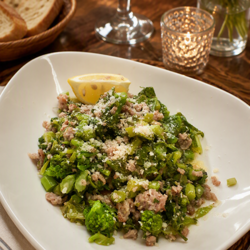 Broccolini with crumbled Italian sausage and grated Parmesan served on a white square plate with lemon.