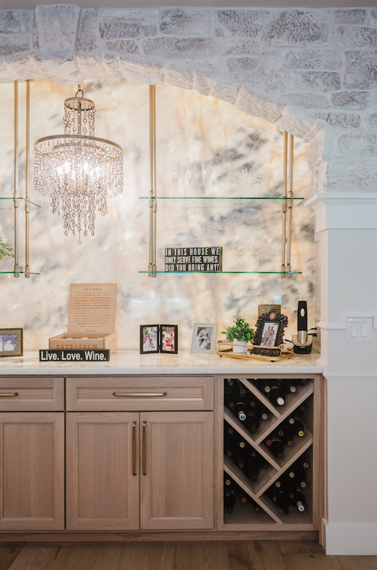 Tuscan-inspired Sea Pines home bar with limestone arch, quartzite backsplash, glass shelves and crystal chandelier