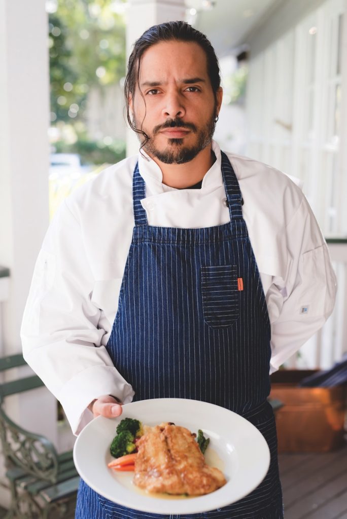 Male chef in a white jacket and dark apron poses in a restaurant setting.