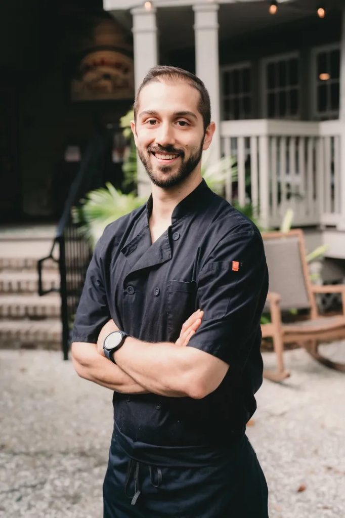 Male chef in a dark chef jacket stands with his arms crossed.