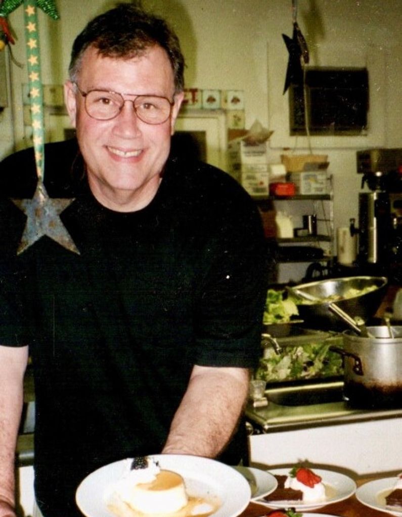 Smiling male chef in black clothing leans forward in a restaurant kitchen.
