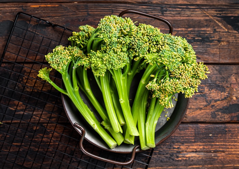 Fresh broccolini stalks with small green florets arranged on a dark wooden tabletop.